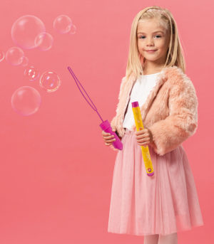 cute and smiling kid holding bottle with soap bubbles on pink ba