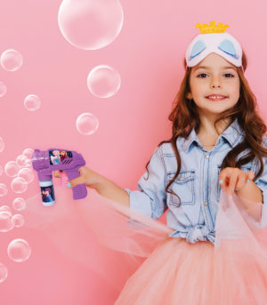Joyful pretty young girl with long brunette hair dancing in tulle skirt isolated on pink background. Amazing cute little princess with mask on head smiling, expressing positivity to camera.